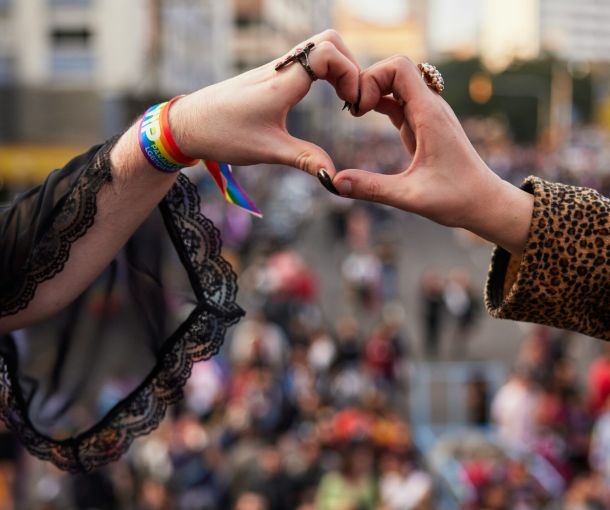 Two people at a Pride event form a heart shape with their hands, celebrating love and unity, symbolizing neuroqueer-resources.