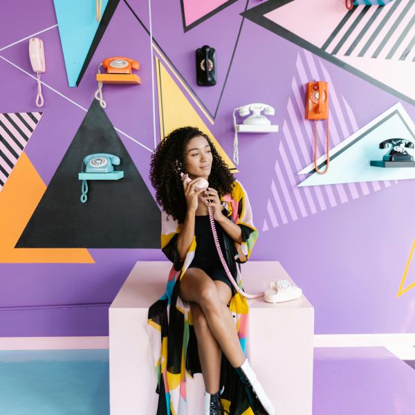 Woman talking on a rotary telephone, sitting in front of a colorful wall displaying multiple telephones.